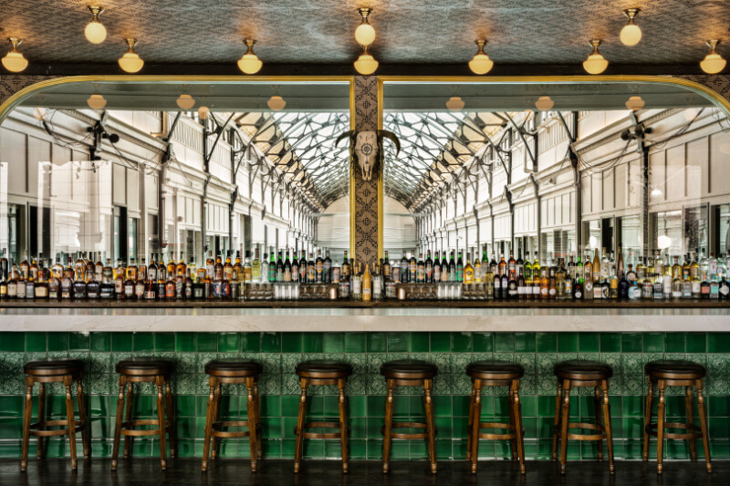 A bar with green tile and seven empty barstools stands in front of a large mirror reflecting a bright, glass-roofed arcade—a stylish setting typical of new Nashville restaurants. Bottles are neatly arranged on the countertop.
